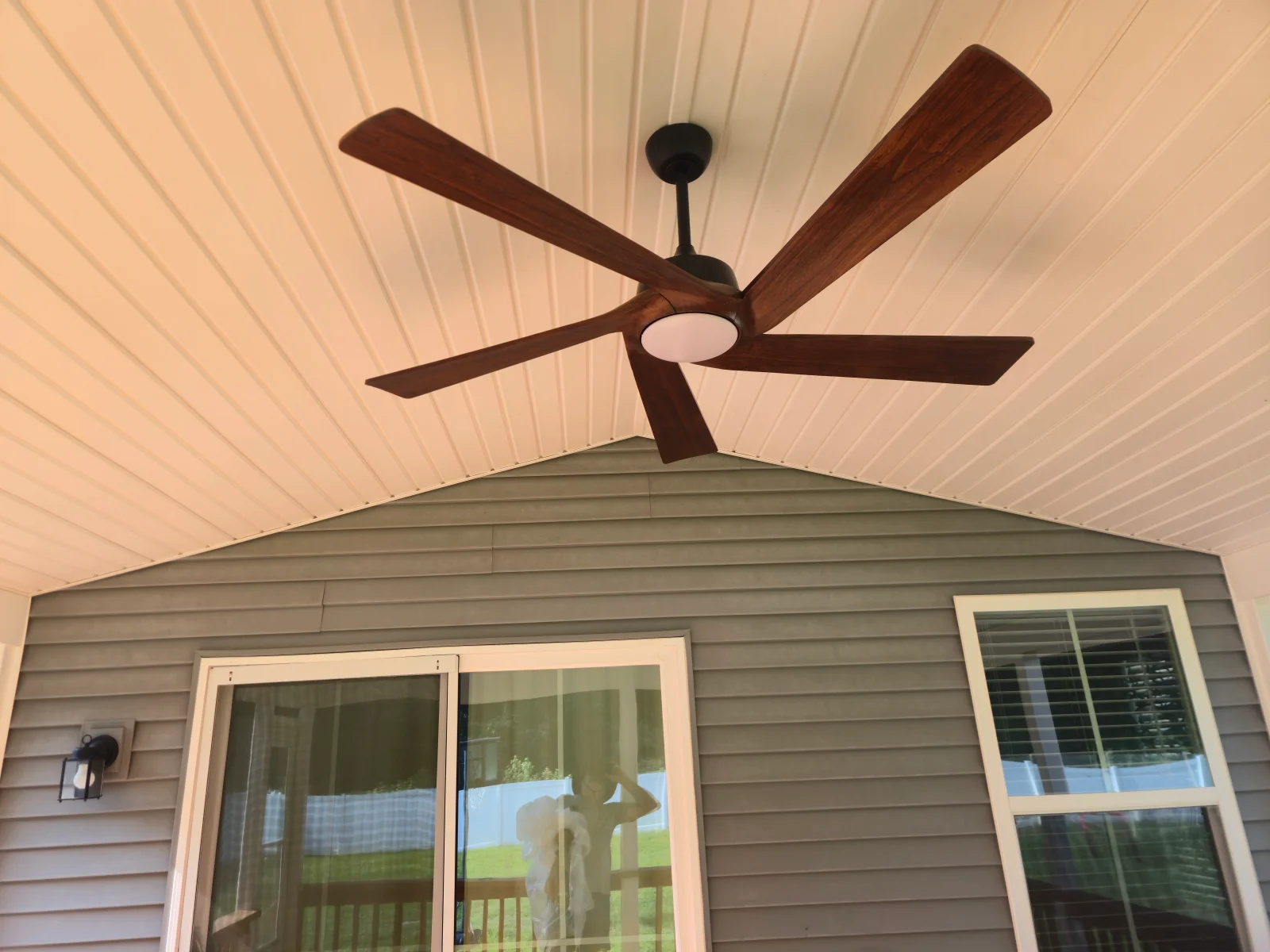 Five-blade wood-finish ceiling fan with frosted light kit on white beadboard porch ceiling above gray vinyl siding