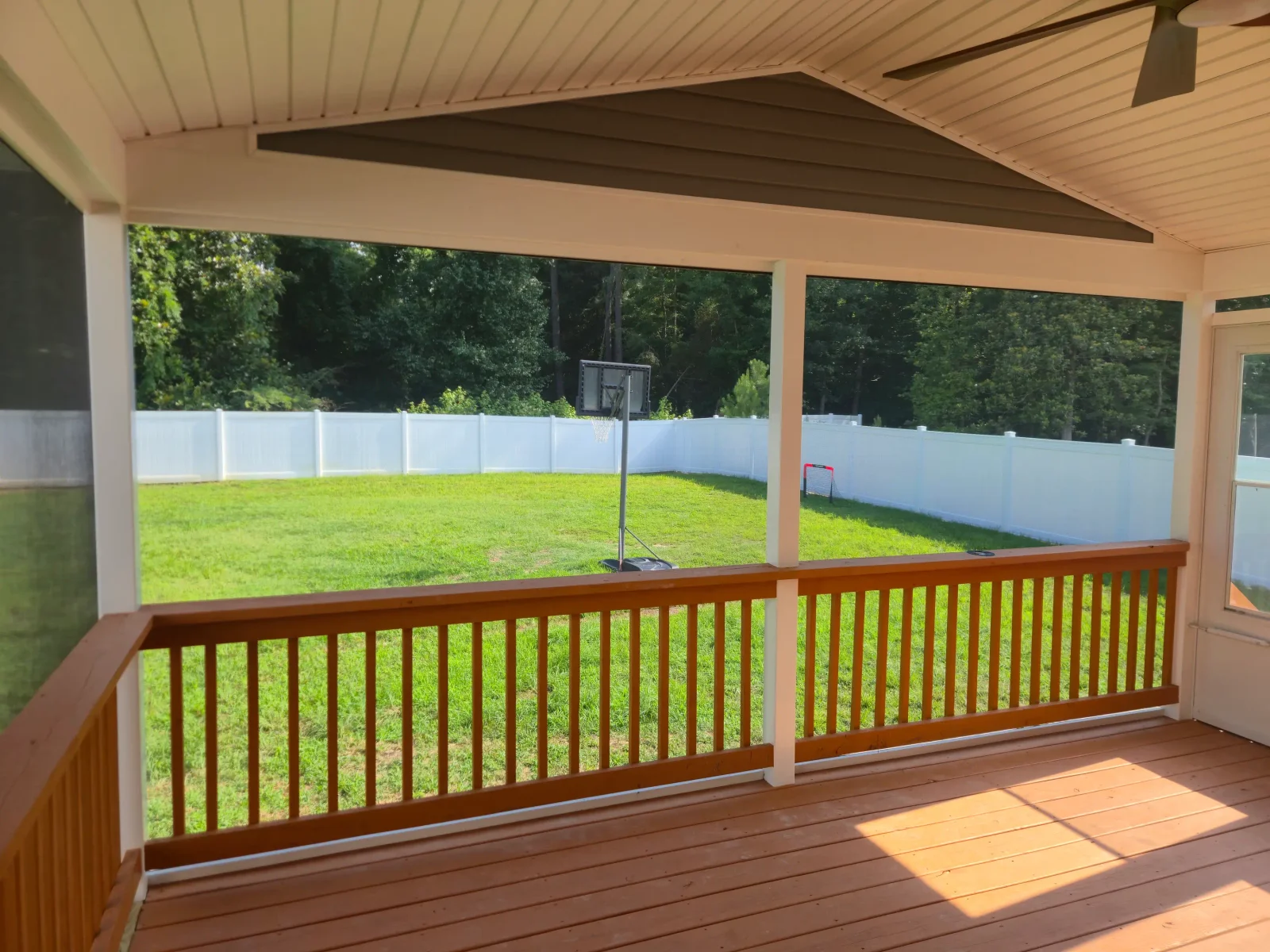 Covered deck with stained wood railing, white beadboard ceiling, and white vinyl fence in backyard
