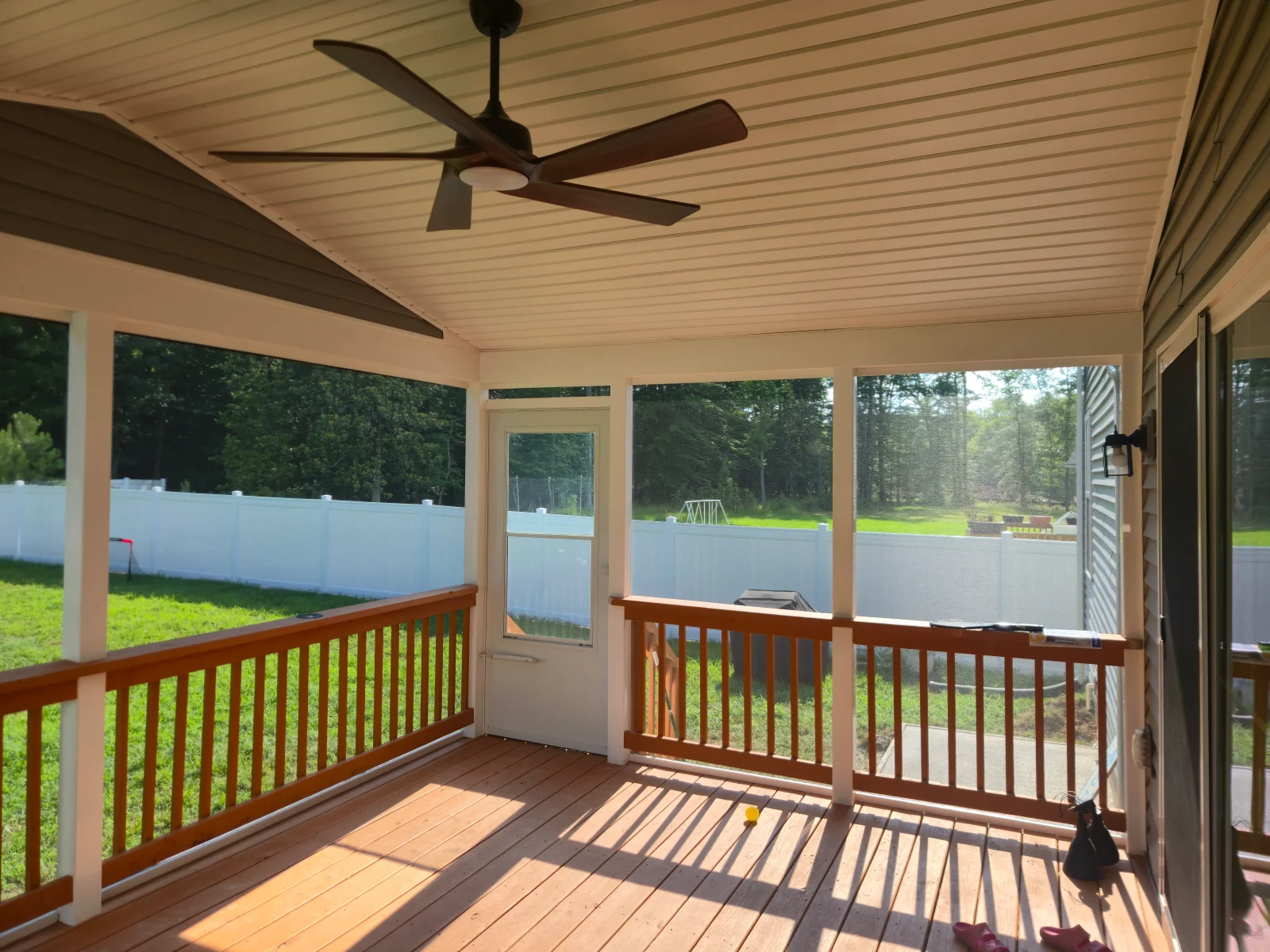 Screened porch with stained wood deck, reddish-brown railings, white posts, tongue-and-groove ceiling, and ceiling fan