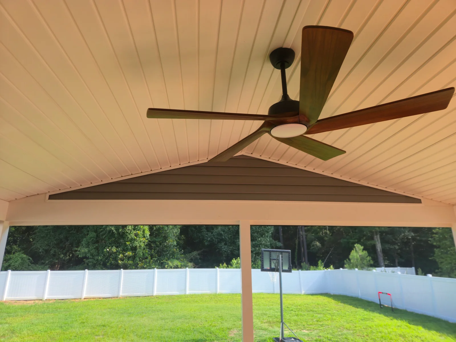 Covered patio with white beaded ceiling, brown gable siding, wood-tone ceiling fan, and white vinyl fence