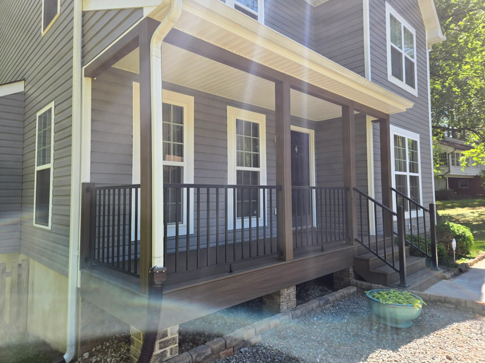 Covered front porch with dark composite decking, brown square posts, black metal railing, and cream beadboard ceiling