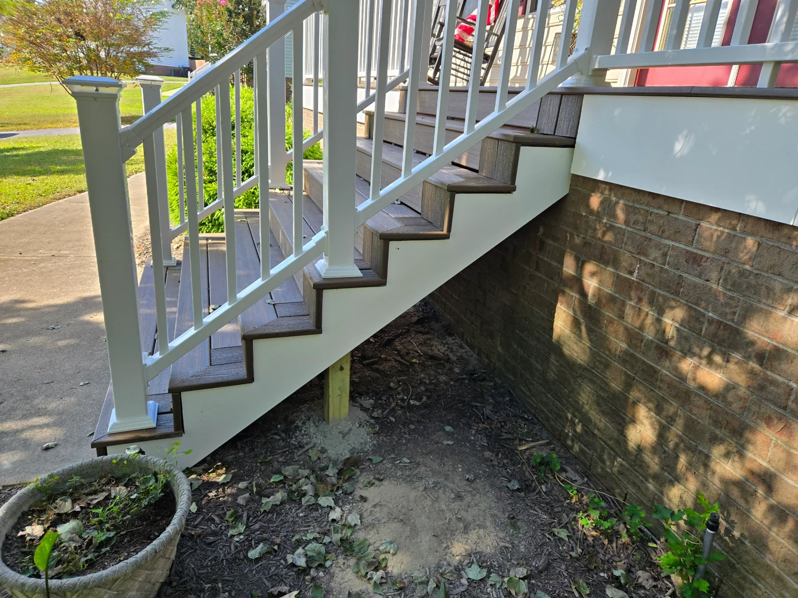 Brown composite deck stairs with white vinyl railing, square balusters, and white stringer skirt beside brick wall