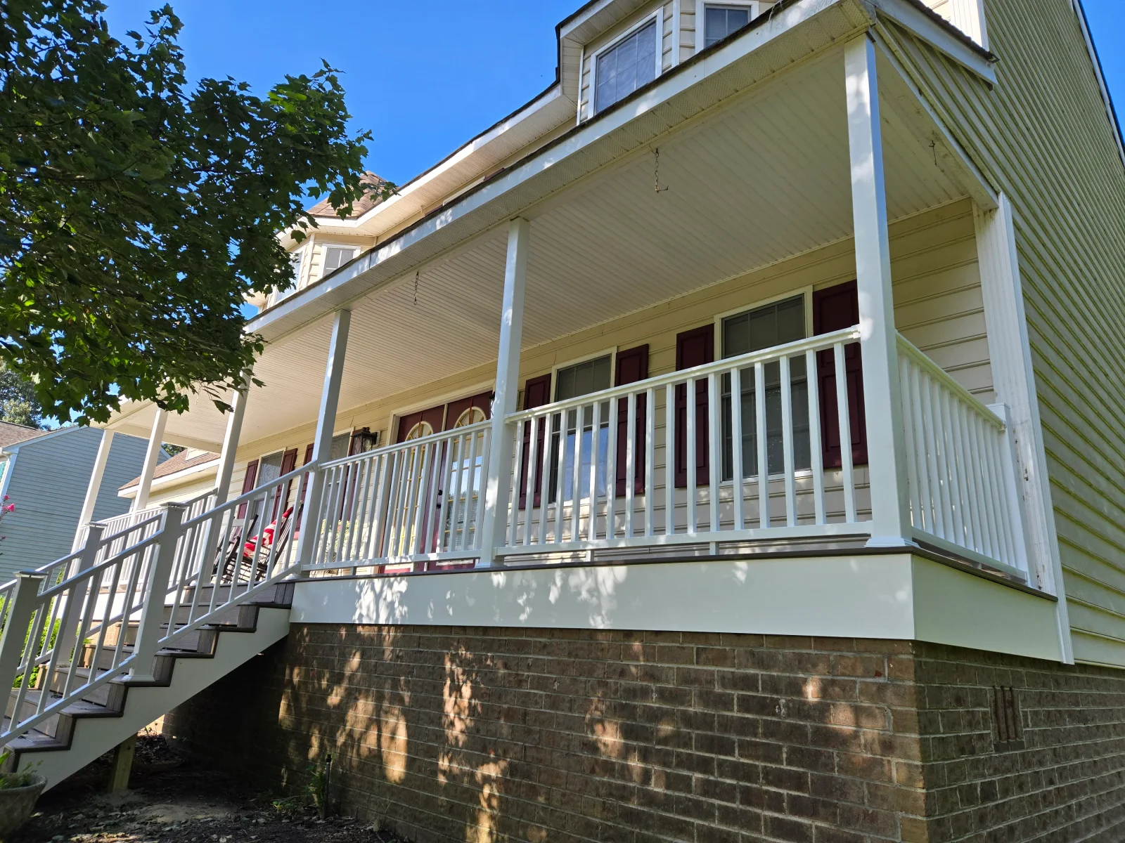 Covered porch with white square columns, white railing, and dark composite steps beside brick foundation and cream vinyl siding