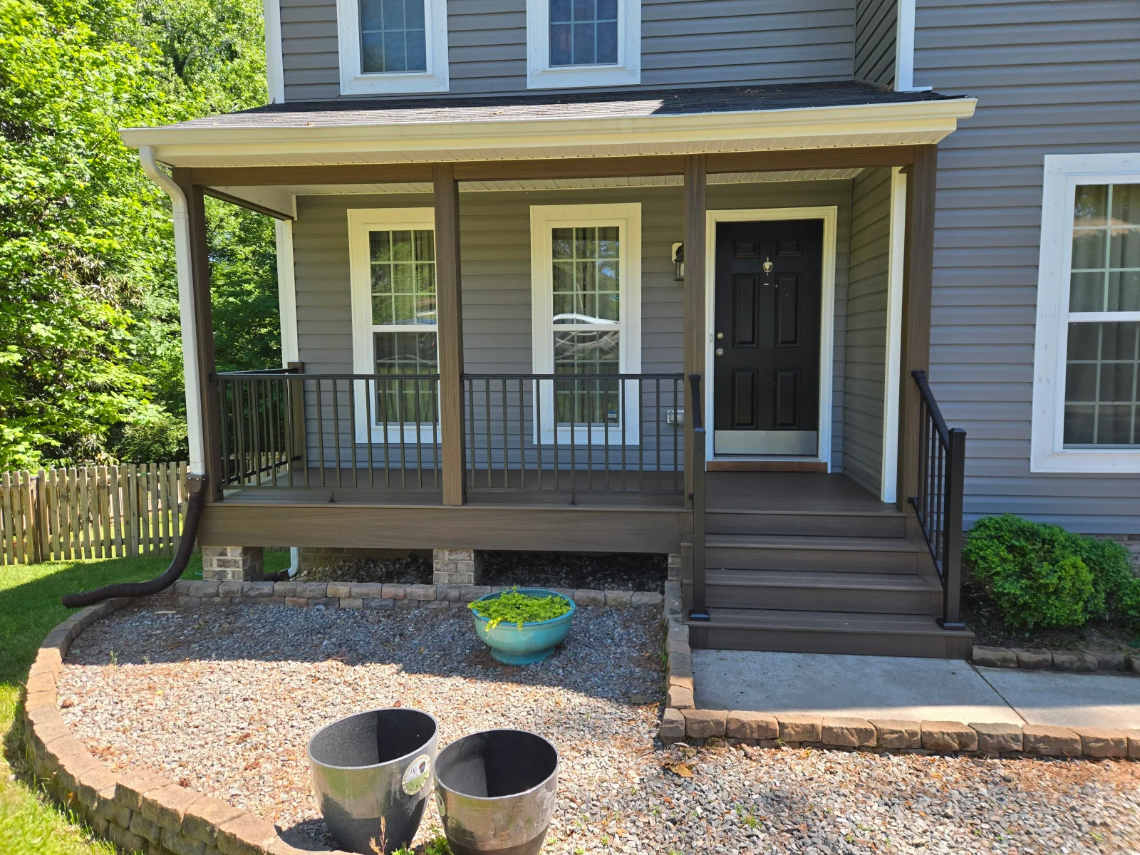 Covered front porch with brown composite decking, black metal railings, and wood-grain posts beside gray vinyl siding