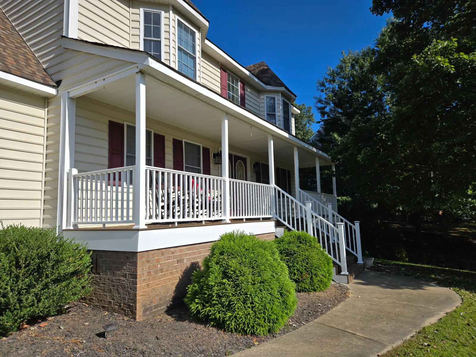 Covered front porch with white columns, vertical baluster railings, tan decking, and white stair rails beside cream vinyl siding